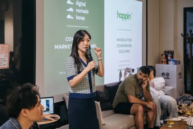 小聚上，一位講者拿著麥克風站在投影幕前分享，旁邊有人坐著聆聽